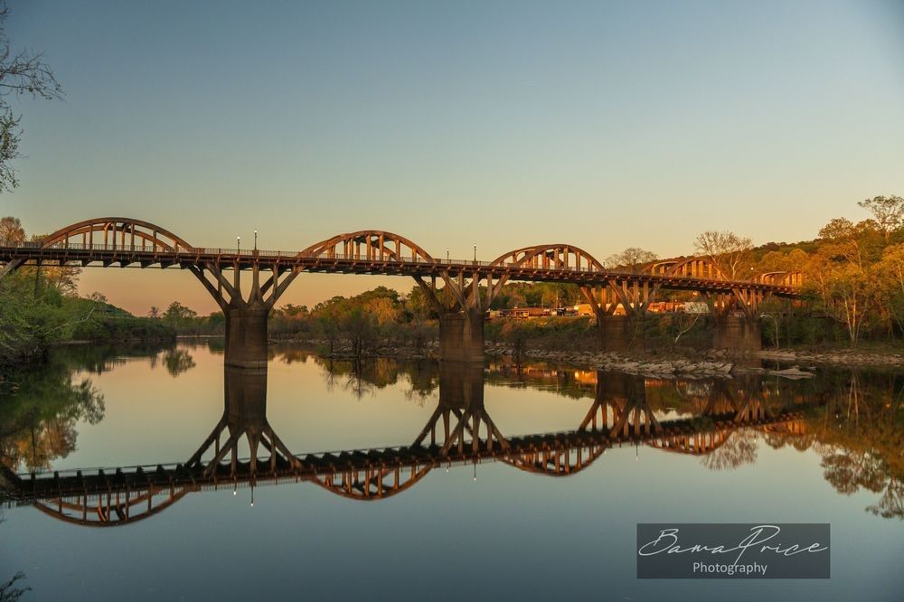 A wide landscape photograph of the historic Bibb Graves Bridge in Wetumpka, Alabama, at sunset. The concrete arches of the bridge are brilliantly illuminated by a warm, golden twilight glow, which is perfectly mirrored in the glassy, still surface of the Coosa River below. The sky fades from a soft gold near the horizon to a clear blue above. Rich green trees line the riverbank. In the bottom right corner, a dark translucent watermark reads "Bama Price Photography."