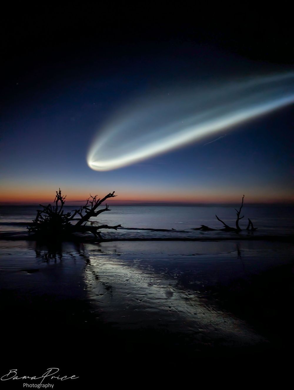 “Uninvited Guest”: A vertical fine art photograph taken at 5:30 a.m. on Driftwood Beach, Jekyll Island, Georgia. A glowing SpaceX Falcon 9 exhaust plume sweeps in a wide luminous arc across a deep blue twilight sky, fanning outward from the upper left to the right side of the frame. Along the horizon, a warm band of orange and red marks the last of the pre-dawn light. In the middle ground, the dark silhouettes of weathered driftwood trees rise from the shoreline. The wet sand in the foreground reflects the glow of the plume above. A Bama Price Photography watermark appears in the bottom left corner.
