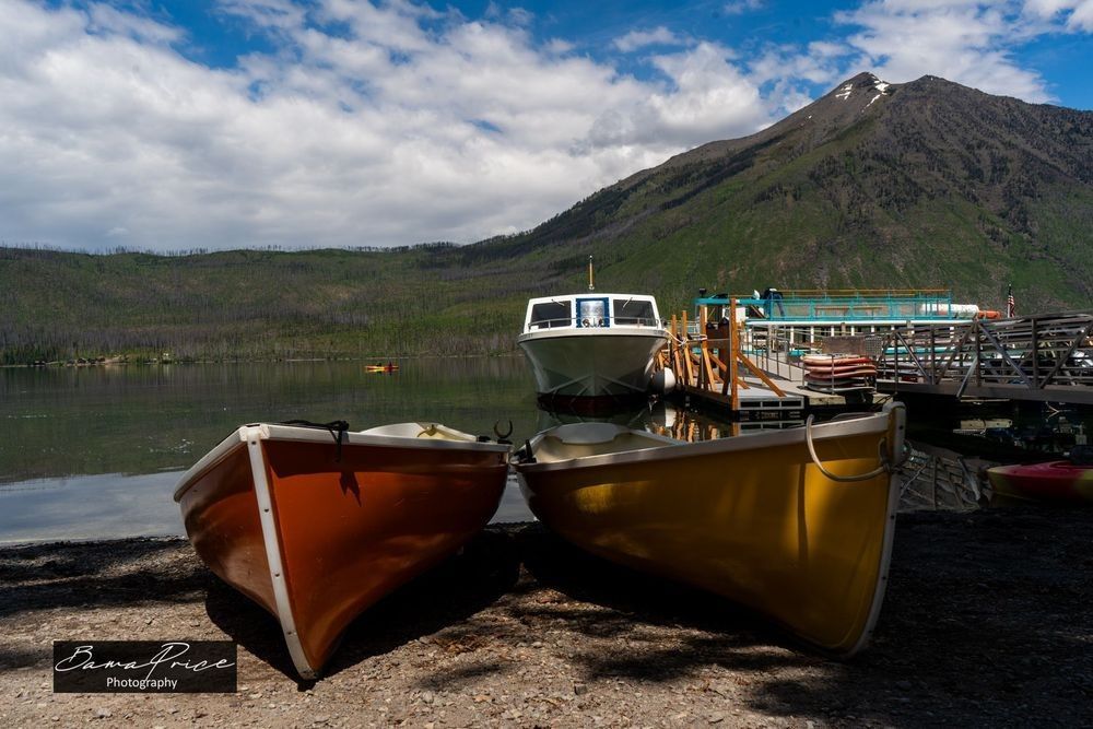 “Lake McDonald’s Invitation”: A horizontal fine art photograph of Lake McDonald at Glacier National Park, Montana, captured on a cloudy June morning. A row of brightly colored boats — reds, yellows, and other vivid hues — are moored along a dock in the foreground. The calm, glassy surface of the lake stretches into the middle ground, reflecting the soft overcast sky. In the background, a snow-capped mountain peak rises above a dark green tree line. A Bama Price Photography watermark appears on the image.