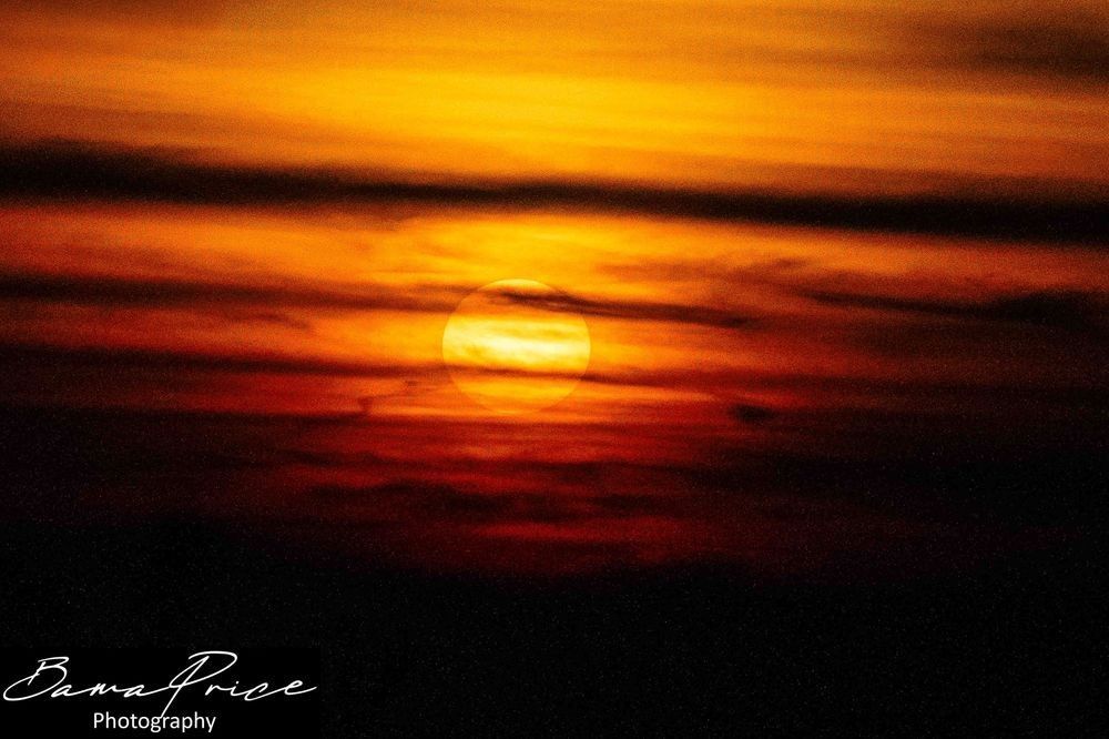 A dramatic, tightly cropped telephoto landscape photograph of a sunset captured from the Bald Rock overlook at Cheaha State Park, Alabama. A massive, glowing yellow sun is partially obscured by heavy, dark, horizontal bands of clouds slicing across the frame. The sky transitions from a brilliant golden-yellow at the top into a deep, fiery orange and dark crimson red at the bottom. In the bottom left corner, the white "Bama Price Photography" watermark is clearly visible.