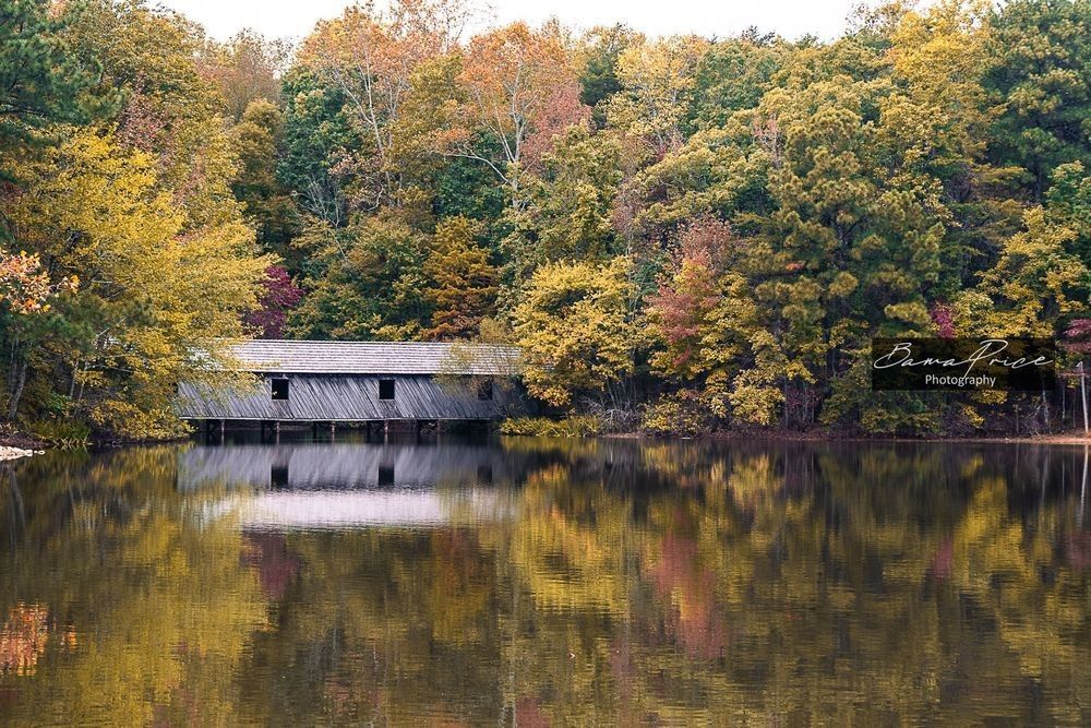 Cambron Covered Bridge surrounded by green, yellow, orange and red autumn foliage reflected in the still water at Madison County Nature Trail in Alabama.