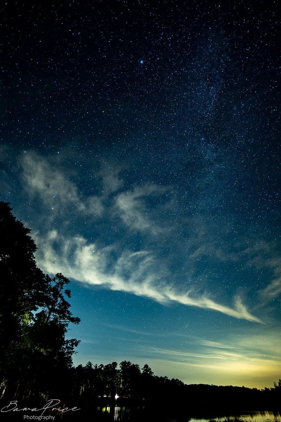 Stars and clouds above a silhouetted tree line and dark water at Payne Lake, Alabama, with distant city glow along the horizon.