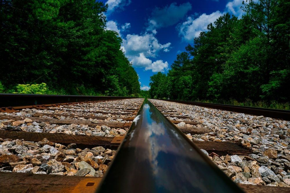 A dramatic, low-angle landscape photograph looking down a straight railroad track in Hoover, Alabama. The camera is positioned directly on the right steel rail, which is polished and perfectly reflects the vibrant blue sky and puffy white clouds above. The wooden ties and gray gravel ballast stone stretch toward a distant horizon line, framed on both sides by dense, rich green summer trees. In the bottom right corner, a dark translucent watermark reads "Bama Price Photography."