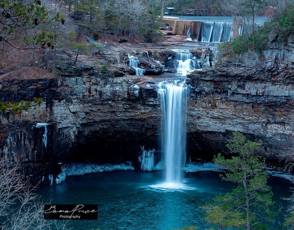 A horizontal landscape photograph of DeSoto Falls near Mentone, Alabama, captured during a winter hard freeze. The multi-tiered waterfall cascades down into a deep teal plunge pool. Large icicles hang from the layered rock cliffs surrounding the lower falls, and white ice chunks line the edges of the pool. The surrounding trees are bare and leafless, showcasing the raw, gray canyon architecture under soft, overcast light.