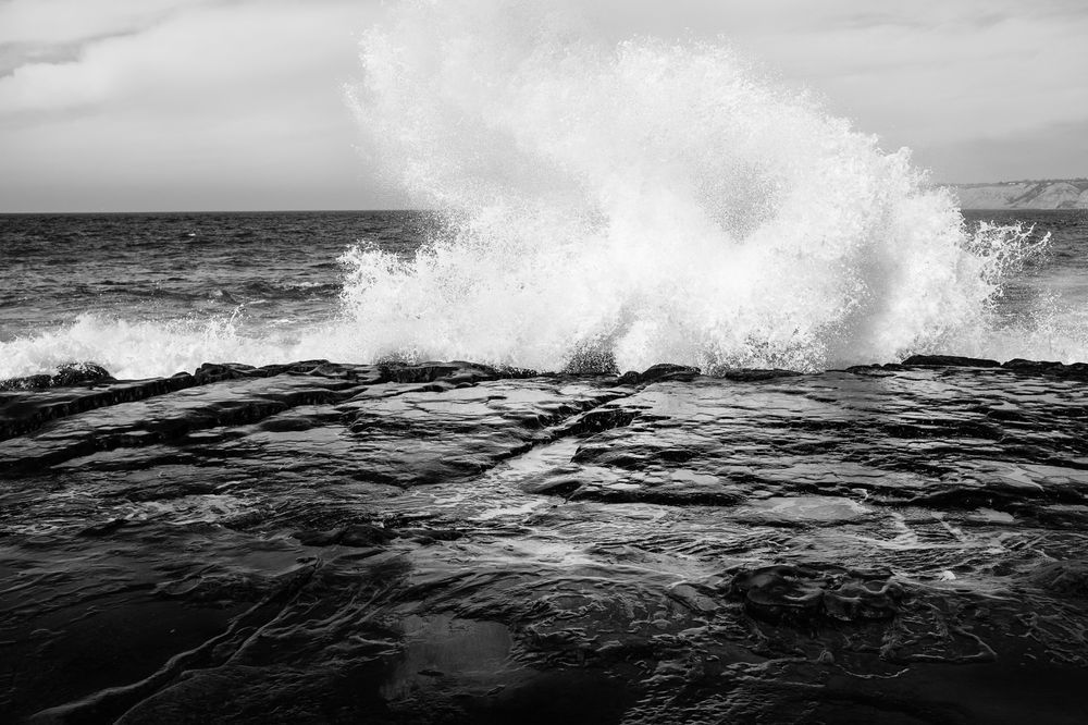 A black and white photograph of a wave crashing against the shore
