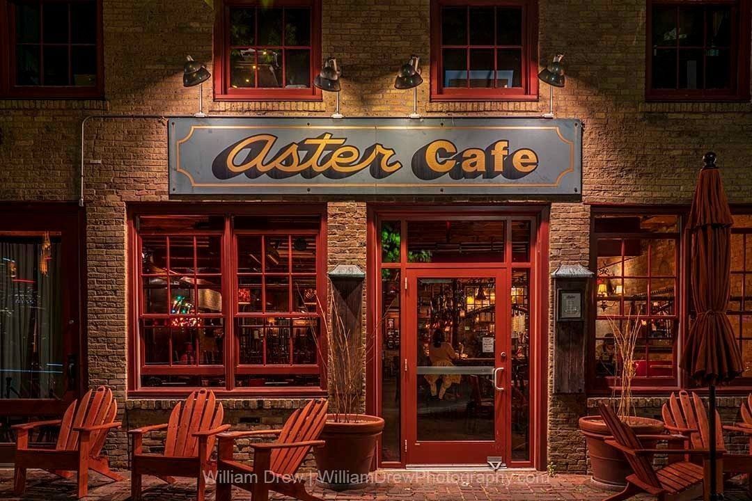 A Night at Aster Café — Minneapolis fine art photography print by William Drew, showing the warmly lit exterior of Aster Café at night with red-trimmed windows, the iconic sign glowing in amber, and Adirondack chairs on the brick patio.