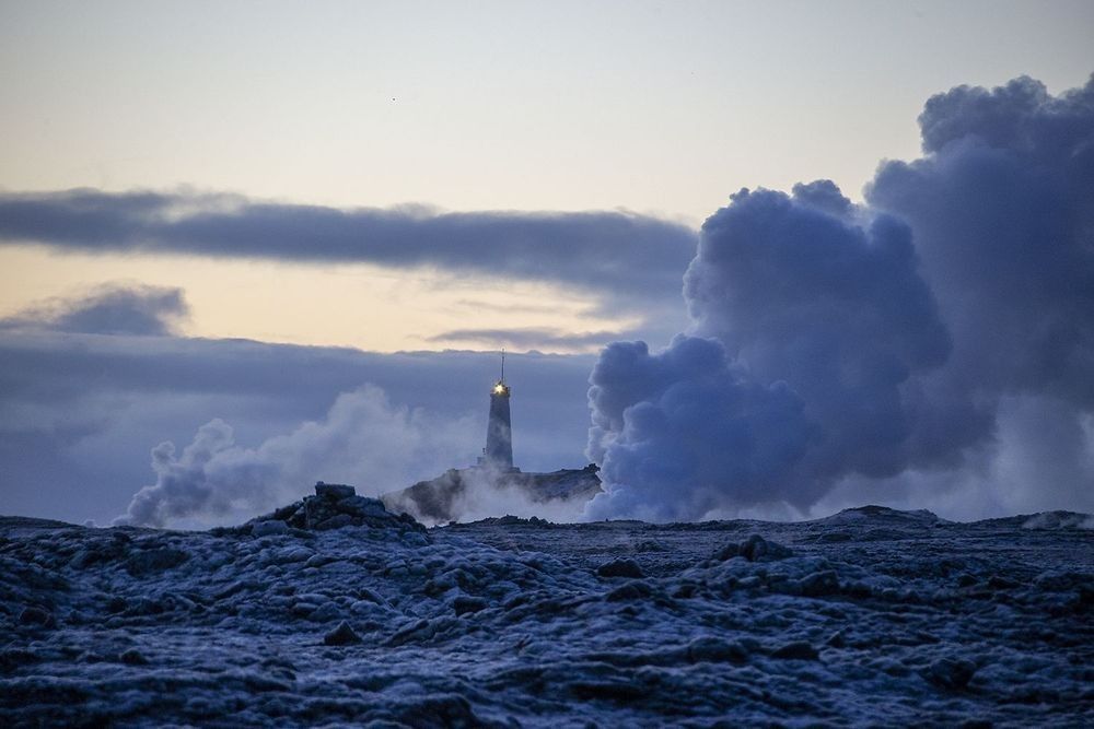 Geothermal steam rising across a volcanic landscape in Iceland at dawn with lighthouse beacon shining through thermal clouds by Robbie George