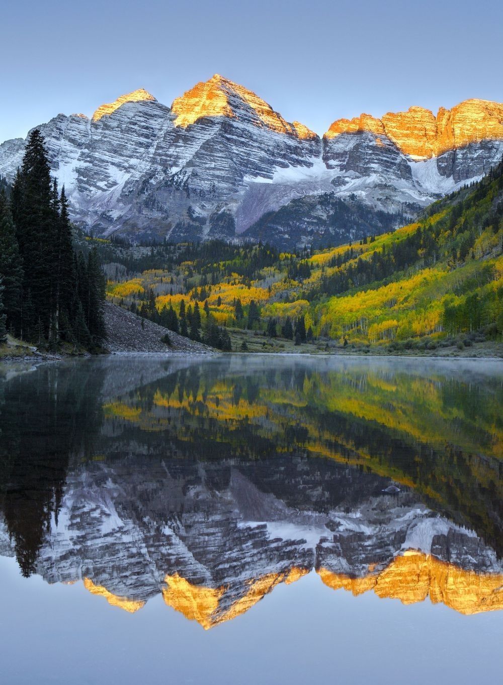 Snow-covered Maroon Bells illuminated by sunrise above an alpine forest and reflective lake, showing the atmosphere, water, ice, rock, vegetation, and solar energy interacting within a mountain climate system.