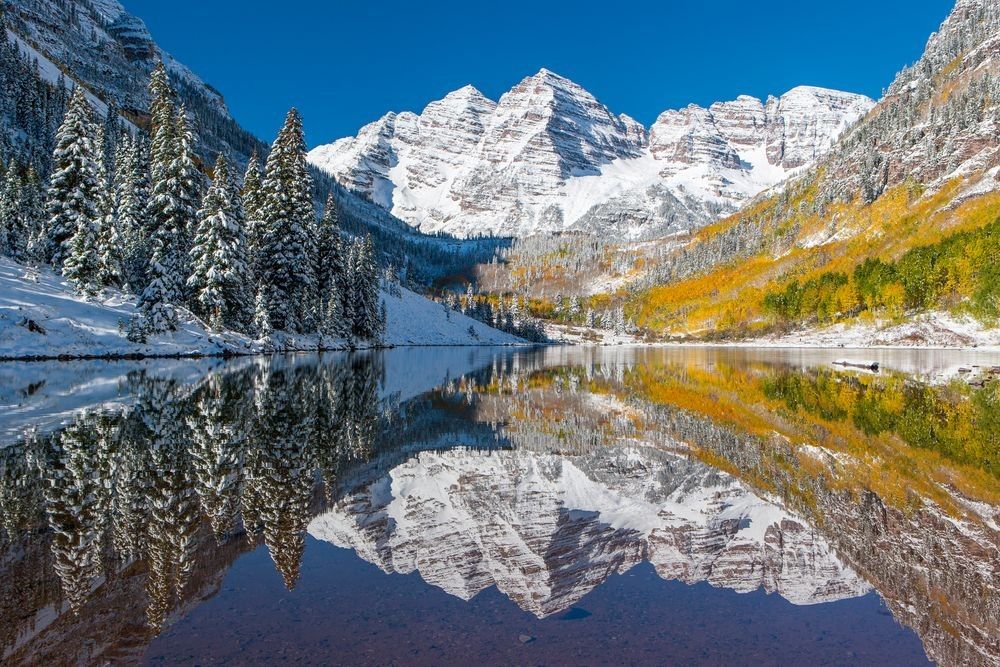 Maroon Bells reflected in alpine lake surrounded by forest and mountain landscape in Colorado photographed by Robbie George