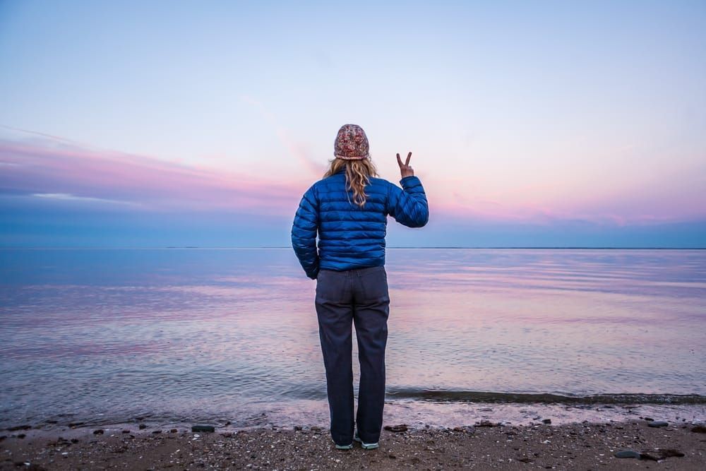 A woman standing at waters edge making a peace sign with her hand / fingers.