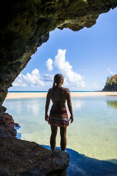 A woman standing at the entrance to a cave looking out over a pool of water to a beach and a ocean beyond.  Hanakapiai Beach, Na Pali Coast of Kauai, Hawaii