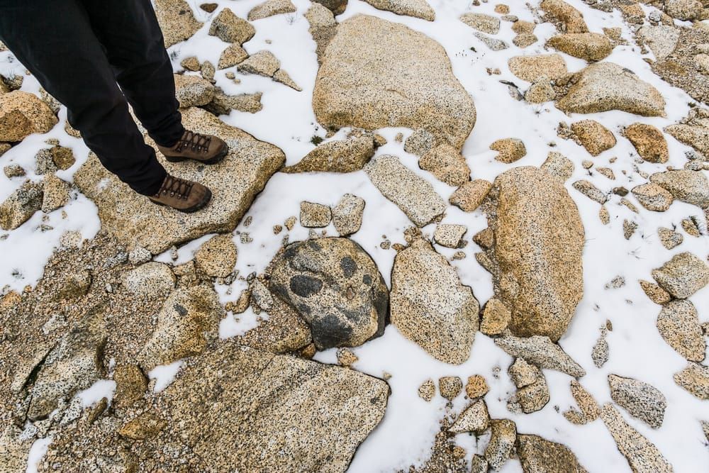 A persons legs and boots standing on a talus and snow slope, Enchantment Lakes Wilderness Area, Washington Cascades, USA.